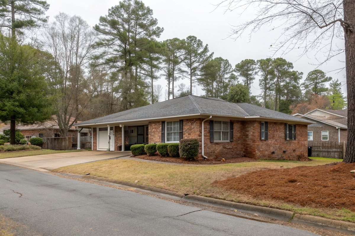 A one-story brick ranch house with a low gray shingle roof, carport and pine straw beds